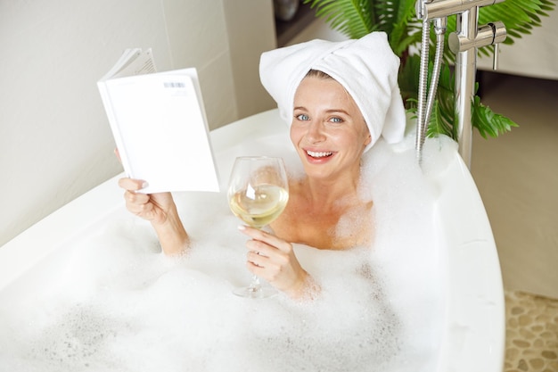 Portrait of pretty smiled young female lying in bath with foam reading and drinking wine