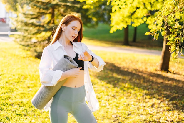 Portrait of pretty smiling redhead young woman checking her smartwatch and holding yoga mat in hand while standing at city park in sunny summer morning