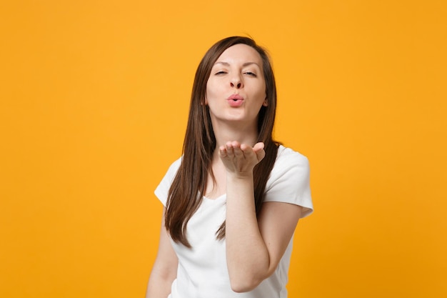 Portrait of pretty tender young woman in white casual clothes looking camera, blowing sending air kiss isolated on bright yellow orange wall background. People lifestyle concept. Mock up copy space.