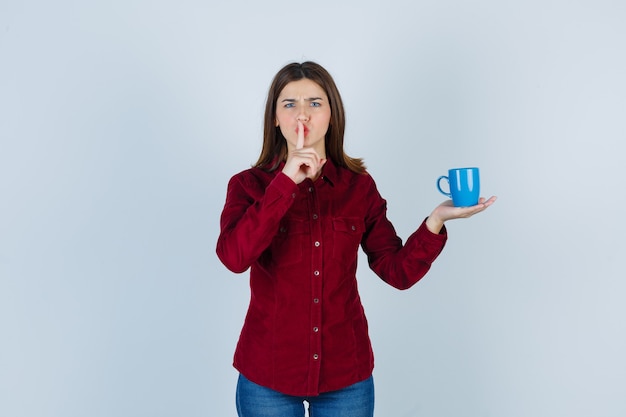 Portrait of pretty woman holding cup on palm, showing silence gesture in burgundy blouse and looking disturbed