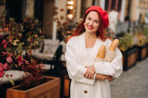 Portrait of a pretty woman in a red beret and a white cardigan with baguettes in her hands strolling through the autumn city