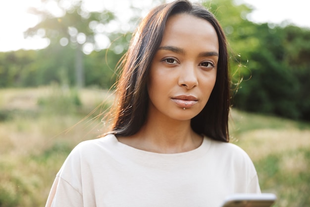 Portrait of pretty woman wearing lip piercing looking at camera and holding smartphone in green park