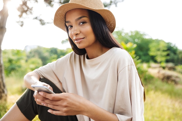 Portrait of pretty woman wearing lip piercing and straw hat holding smartphone while sitting on bench in green park