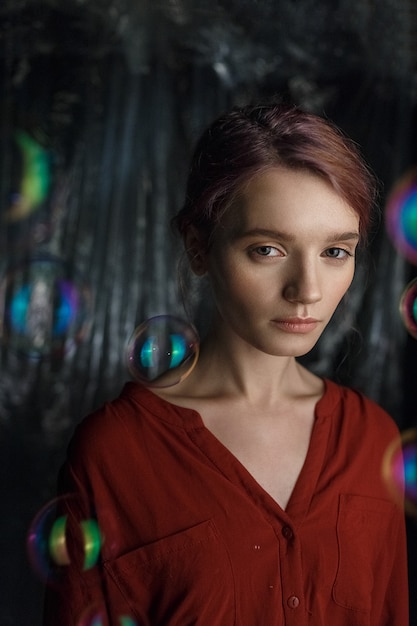 Portrait of pretty young caucasian girl in red shirt. Soap bubbles fly around her head shimmering with rainbow colors.