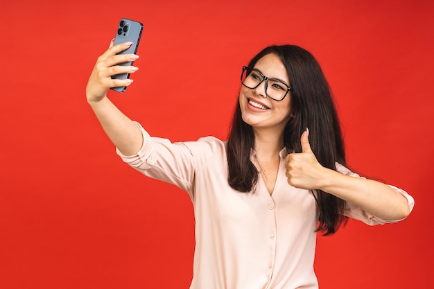 Portrait of pretty young happy woman making selfie on smartphone isolated over red background