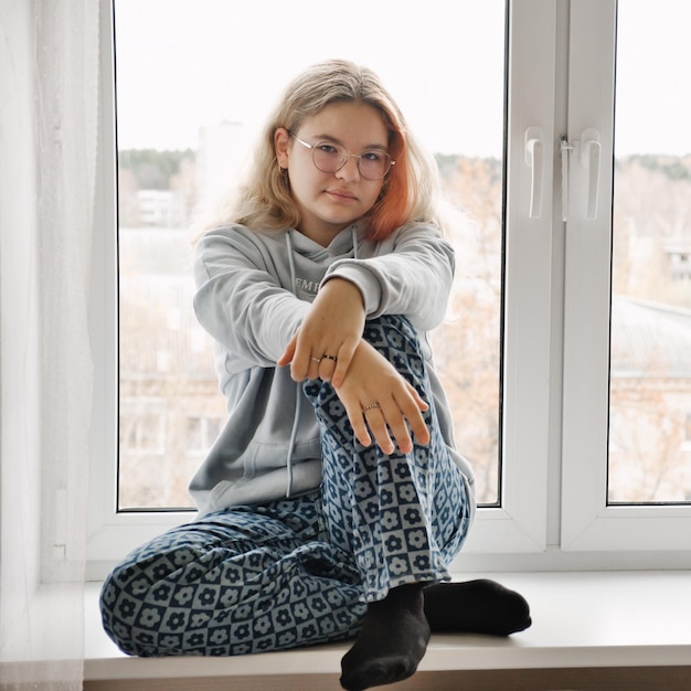 Portrait of the pretty young teenage girl in glasses and casual home clothes sitting on windowsill