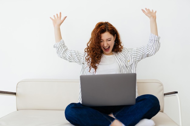 Portrait of a pretty young woman celebrating a success on laptop sit on couch