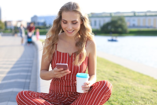 Portrait of pretty young woman sitting on riverbank with legs crossed during summer day, using smartphone.