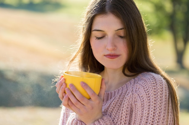 Portrait of pretty young woman in stylish knitted sweater standing on the golden autumn forest on sunny day Time to relax and have some coffee