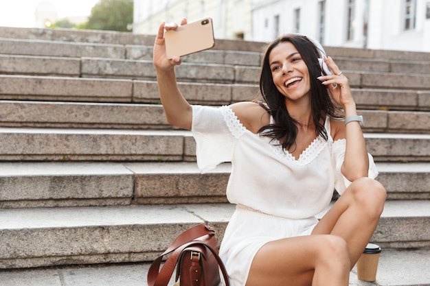 Portrait of pretty young woman wearing headphones taking selfie photo on smartphone while sitting on stairs outdoors