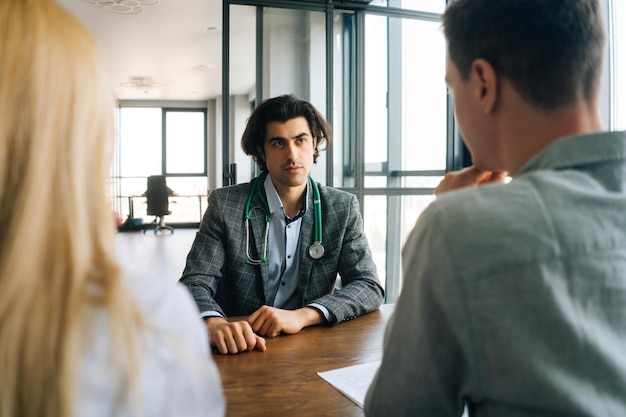 Portrait of professional male doctor with stethoscope talking to unrecognizable man and woman about health during counseling at medical office Serious physician offering advices to young couple