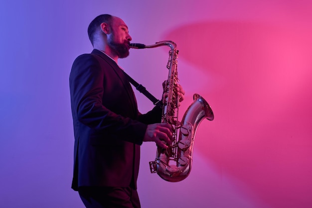 Portrait of professional musician saxophonist man in suit plays jazz music on saxophone, pink background in a photo studio, side view