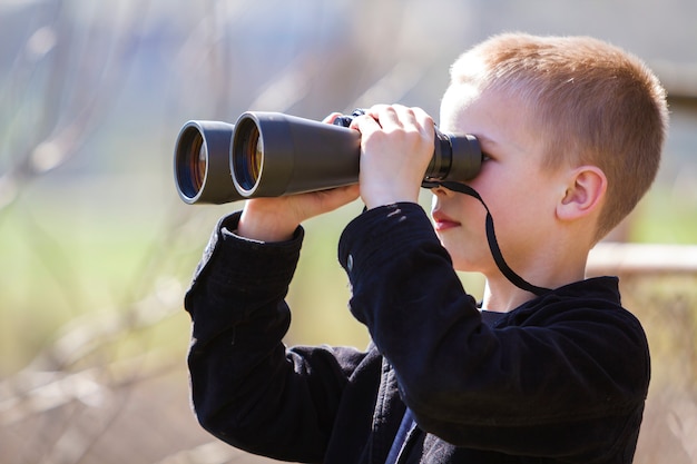 Portrait in profile of little handsome cute blond boy looking thoughtfully through binoculars 