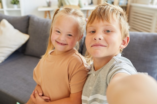 Photo portrait of proud brother taking selfie with cute girl with down syndrome while sitting on sofa at home together, copy space