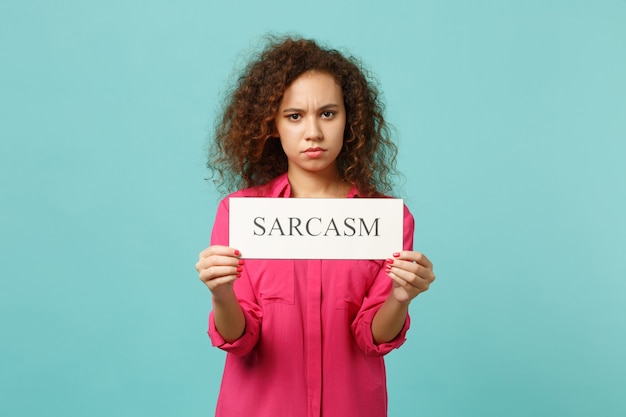 Photo portrait of puzzled african girl in pink casual clothes holding text board sarcasm isolated on blue turquoise wall background in studio. people sincere emotions, lifestyle concept. mock up copy space.