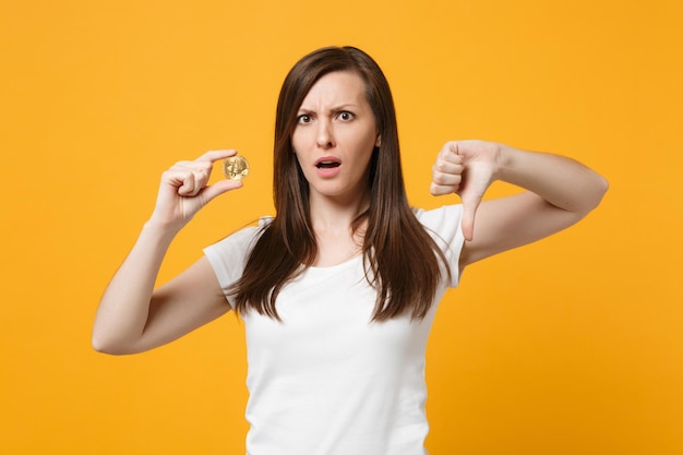 Portrait of puzzled young woman in white casual clothes showing thumb down, hold bitcoin, future currency isolated on yellow orange background in studio. People lifestyle concept. Mock up copy space.