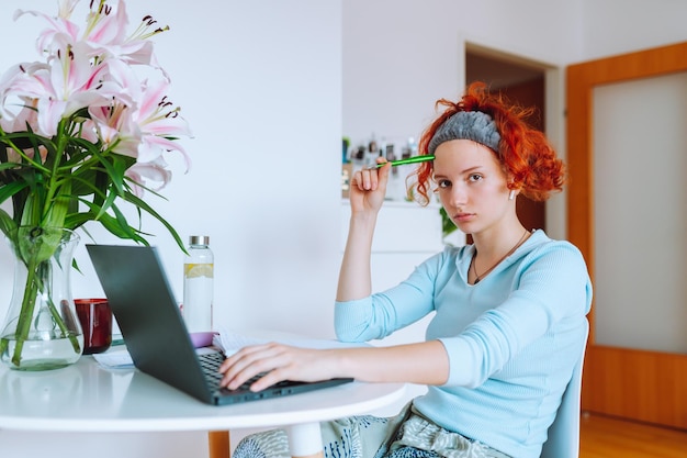 Portrait redhaired teenage girl sitting at table in room using computer