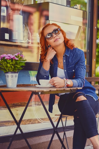 Portrait of redhead female in sunglasses, drinks coffee in a cafe on a street.