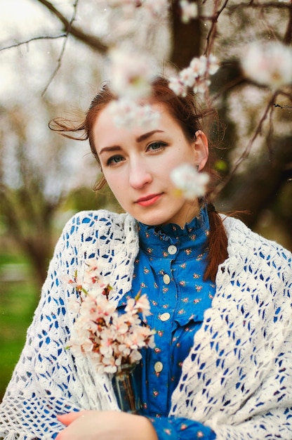 portrait of a redhead young woman in a blue coat and dress in a rainy spring garden of apple trees