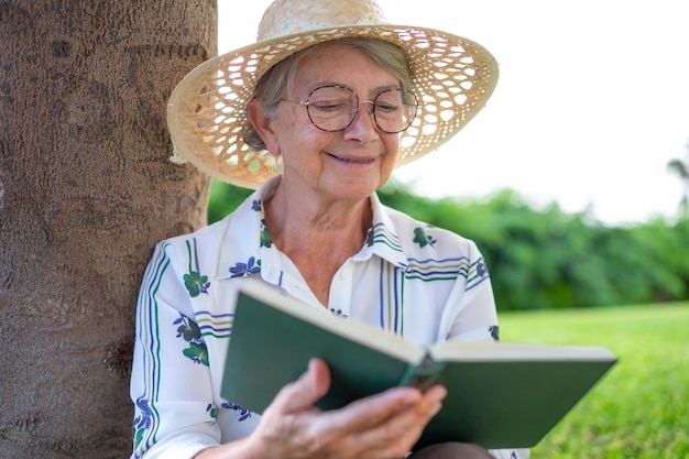 Portrait of relaxed attractive senior woman with hat and eyeglasses sitting on the meadow of public park reading a book caucasian lady enjoying free time and retirement