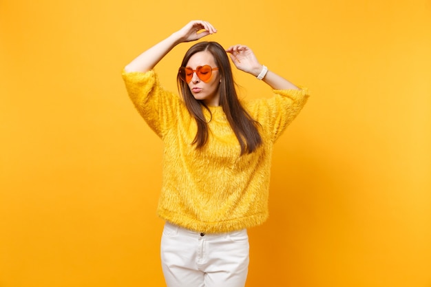 Photo portrait of relaxed young woman in fur sweater, white pants and heart orange glasses rising hands up isolated on bright yellow background. people sincere emotions, lifestyle concept. advertising area.