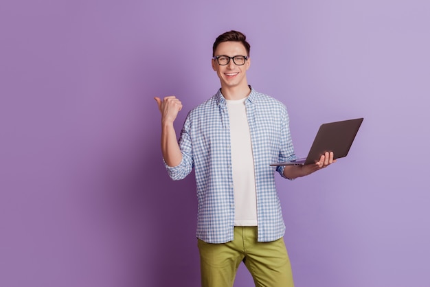 Portrait of reliable freelancer guy hold laptop indicate thumb blank space on violet background