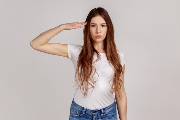 Portrait of responsible serious woman with dark hair saluting commander listening order with obedient expression wearing white Tshirt Indoor studio shot isolated on gray background