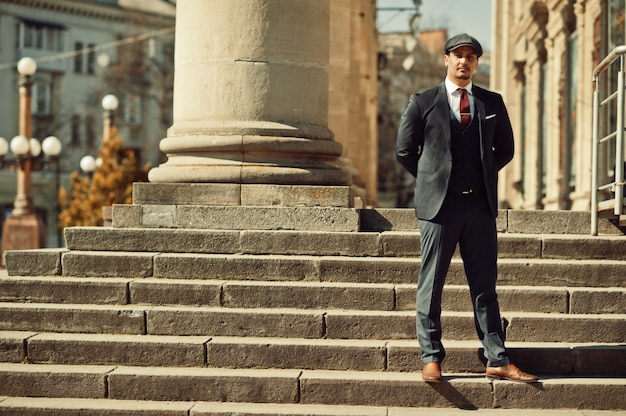 Portrait of retro 1920s english arabian business man wearing dark suit, tie and flat cap near old columns.