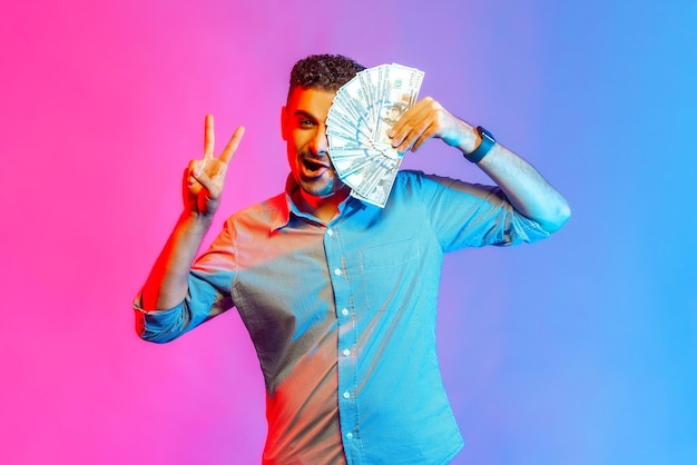 Portrait of rich funny man in shirt covering half of face with dollars, looking at camera showing vi sign, being happy to win lottery. Indoor studio shot isolated on colorful neon light background.