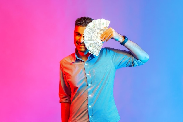 Portrait of rich funny man in shirt covering half of face with dollars looking at camera with toothy smile being happy to win lottery Indoor studio shot isolated on colorful neon light background