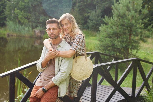 Portrait of romantic adult couple embracing while posing on wooden bridge by lake in rustic countryside scenery