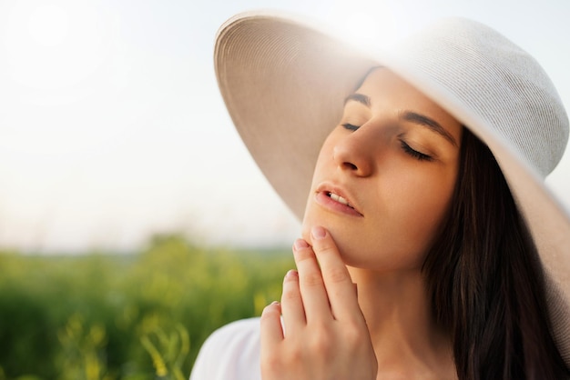 Portrait of a romantic attractive beautiful young woman with a hat and white shirt dreaming with closed eyes on nature field background Beauty