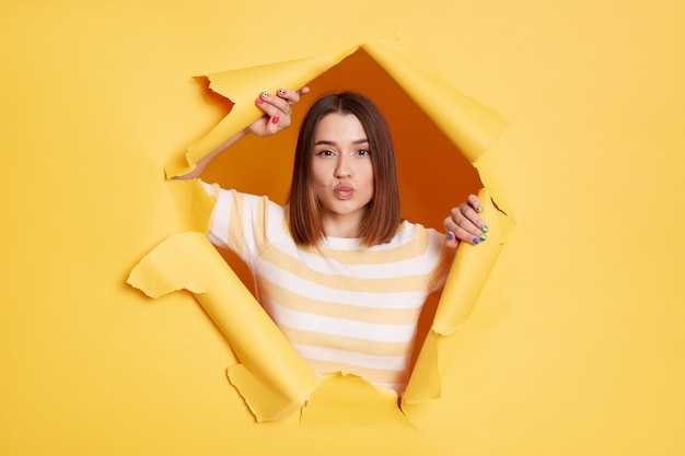 Portrait of romantic lovely adorable woman stands in torn paper hole posing with pout lips and sending air kisses looking through breakthrough of yellow background