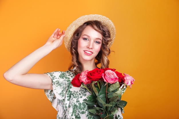 Portrait of romantic young girl in dress and straw hat holding roses bouquet on yellow background