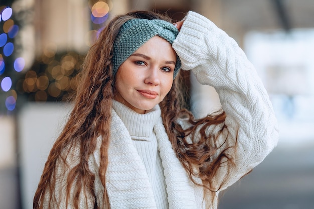 Portrait in the room. A young wonderful girl with long curly hair poses in a soft white look with a raised hand and a slight smile. Casual photo session. The concept of classics.