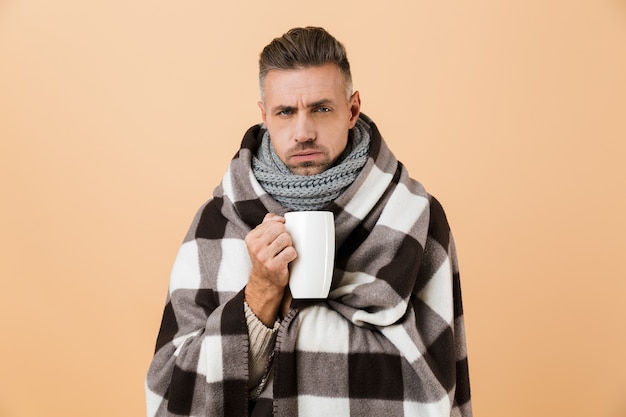 Portrait of a sad sick man wrapped in a blanket standing isolated over beige wall, holding cup with hot tea