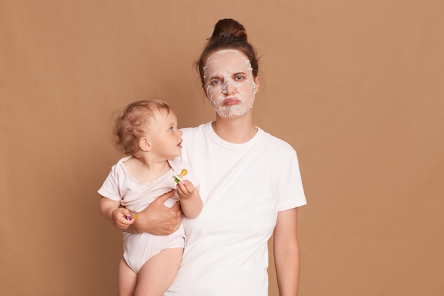 Portrait of sad upset mother holding her little baby doing cosmetic procedures while taking care of her infant daughter posing isolated over brown background