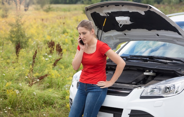 Portrait of sad woman leaning on broken car with open hood and talking by phone