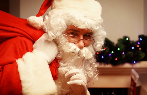 Portrait of Santa Claus with huge red sack keeping forefinger by his mouth and looking at camera