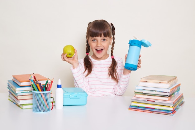 Portrait of satisfied amazed little girl with braids sitting at desk posing isolated over white background being happy to have and snack during break holding apple and bottle with water