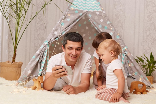 Portrait of satisfied delighted Caucasian man wearing white tshirt with his children posing on floor near peetee tent playing with smart phone expressing happiness having fun together