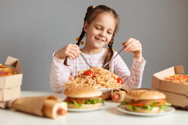 Portrait of satisfied little girl with pigtails sitting at table with big variety of junk food eating pasta with satisfied expression isolated over gray background