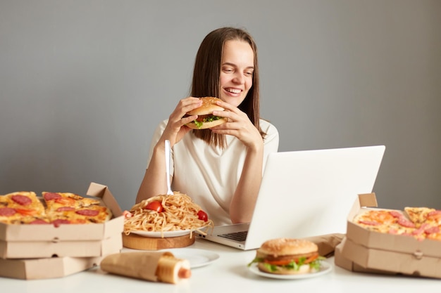 Portrait of satisfied smiling woman watching films or funny video laptop computer eating tasty hamburger wearing white tshirt isolated over gray background