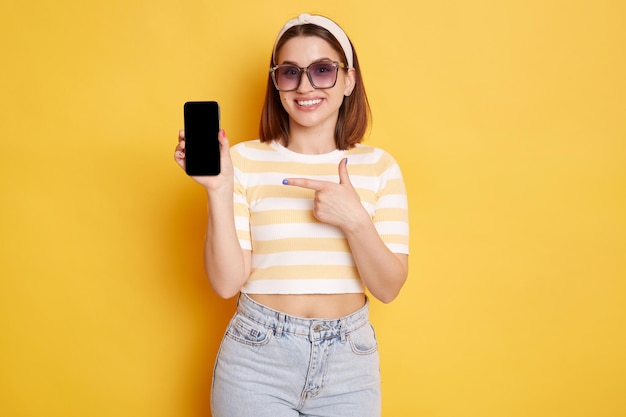 Portrait of satisfied winsome Caucasian woman wearing striped shirt posing isolated over yellow background holding cell phone with advertisement area and pointing at blank screen