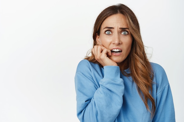 Portrait of scared and alarmed brunette woman, biting fingernails and staring worried at camera, frightened of smth scary, standing over white background. Copy space