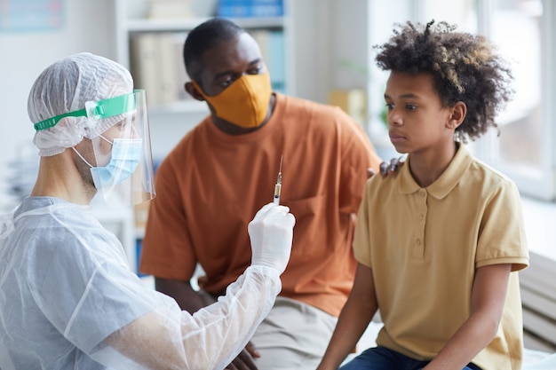 Portrait of scared teenage boy looking at syringe with needle during covid vaccination in clinic with father supporting him