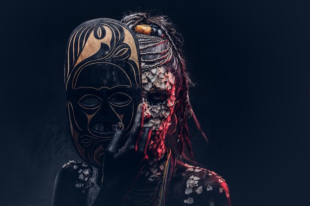 Portrait of a scary African shaman female with a petrified cracked skin and dreadlocks, holds a traditional mask on a dark background. Make-up concept.