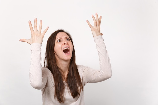 Portrait of screaming young woman in light clothes looking up aside rising, spreading hands isolated on white wall background in studio. People sincere emotions, lifestyle concept. Mock up copy space.