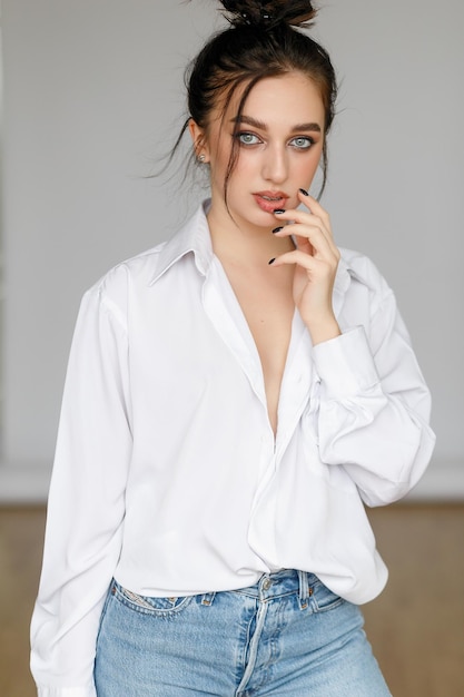 Portrait of seated young woman wearing a bouffant shirt with naked shoulders on light grey background Beautiful brunette girl in jeans and white tshirt smiling