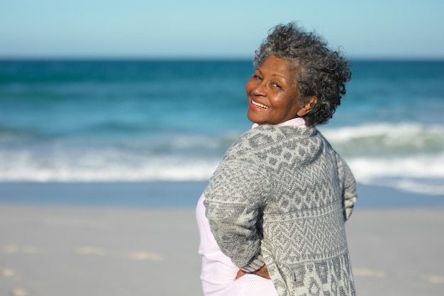 Portrait of a senior African American woman standing on the beach with blue sky and sea in the background, turning around with hands on hips and smiling to camera